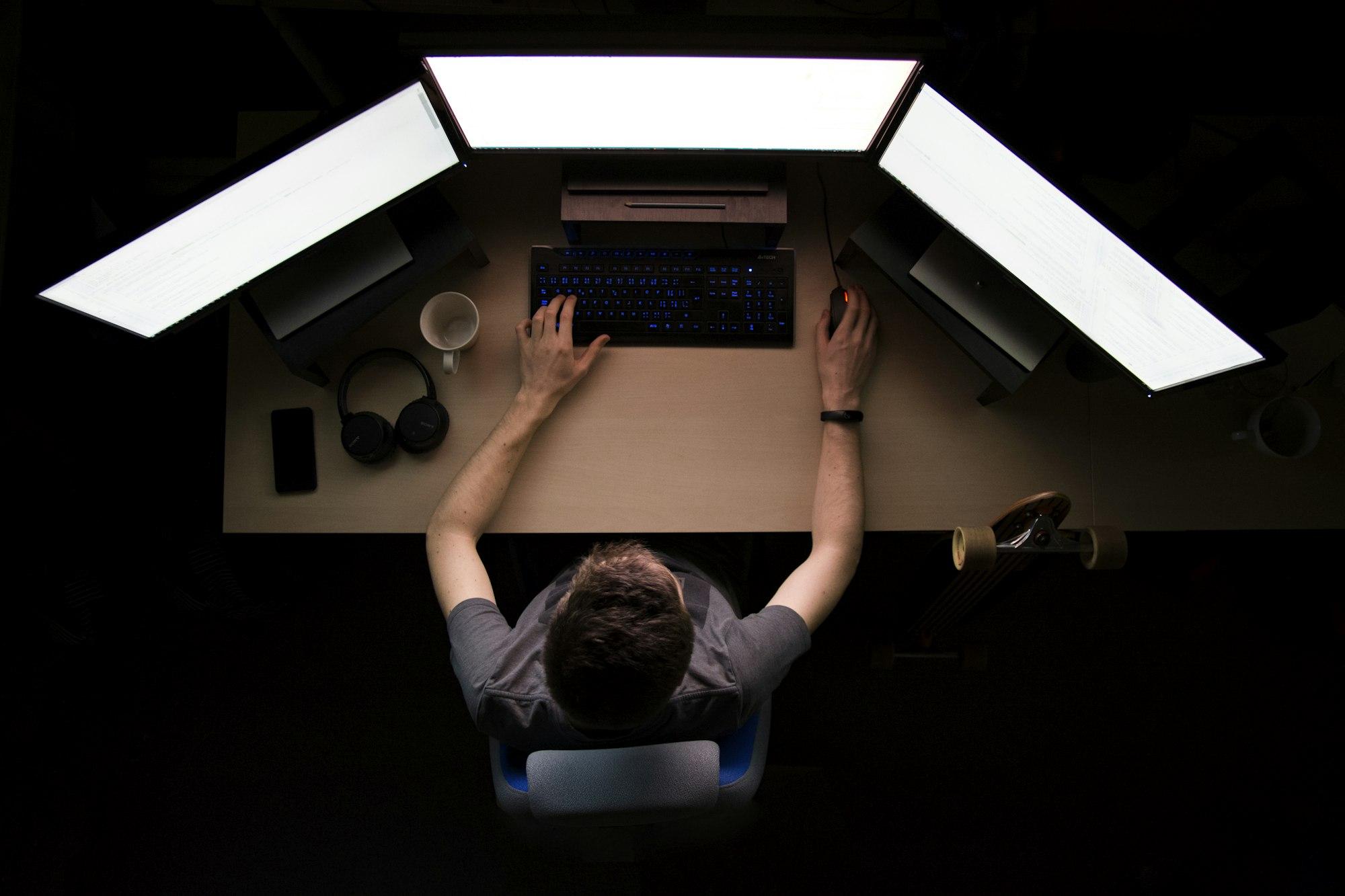 Vista de cima de uma pessoa trabalhando em uma mesa com três monitores, em um ambiente escuro